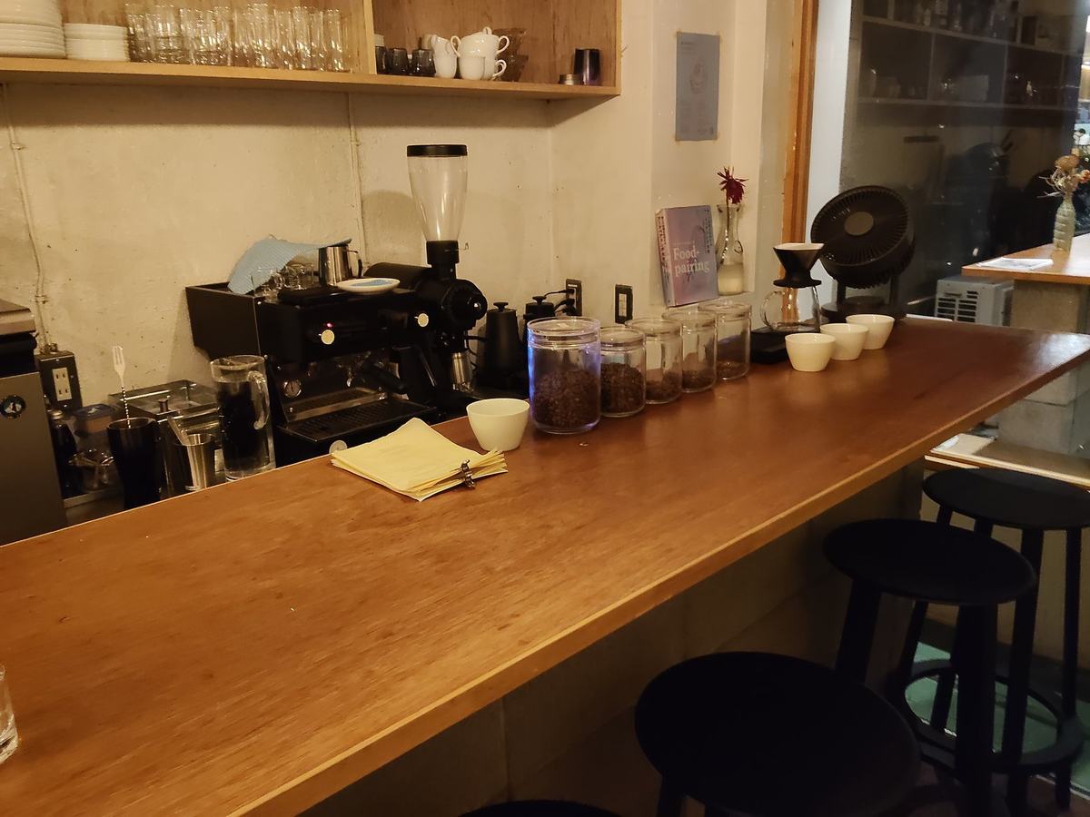 深夜喫茶 幻想の店内。木の一枚板のカウンターが奥へ伸び、黒い丸いスツールが並ぶ。壁の棚にグラスと豆の瓶。