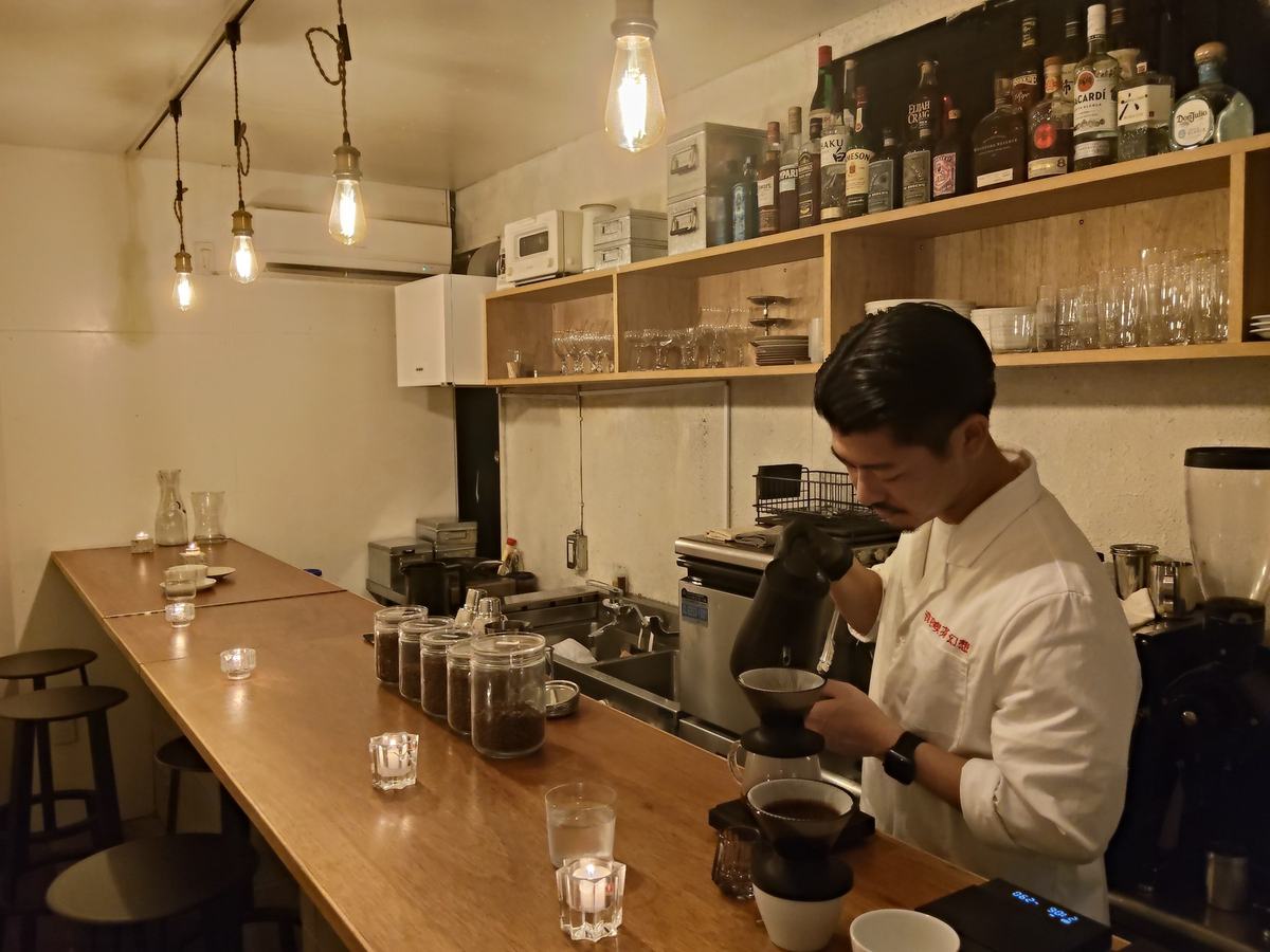 端から端まで見渡した深夜喫茶 幻想のカウンター。天井に電球が並び、奥で白い上着の男性がコーヒーを淹れている。
