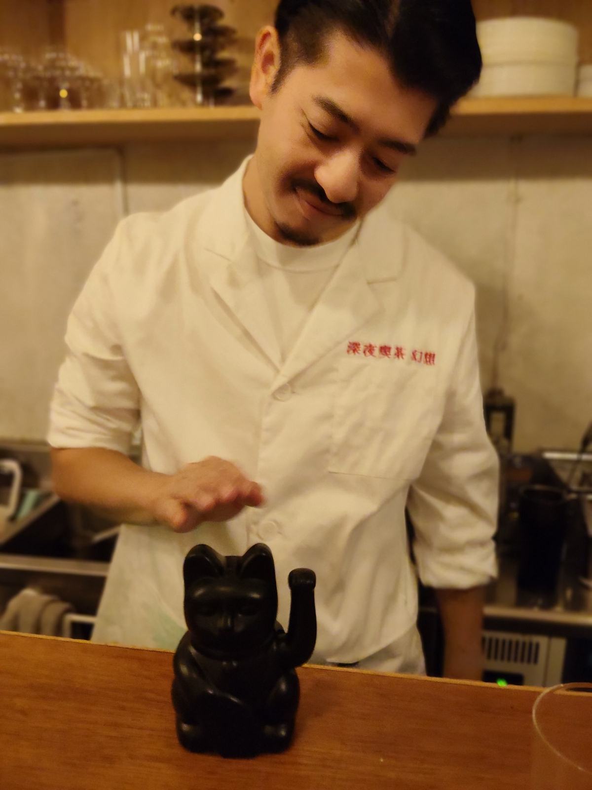 カウンターに置かれた黒い招き猫。その後ろで白い上着の男性が、猫の頭の上に手をかざしている。