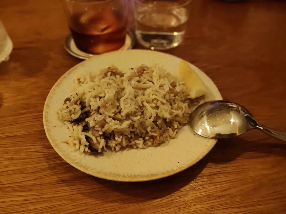 野菜ビリヤニ。白い皿に盛った茶色い炊き込みご飯に、櫛切りのレモンとスプーンが添えてある。