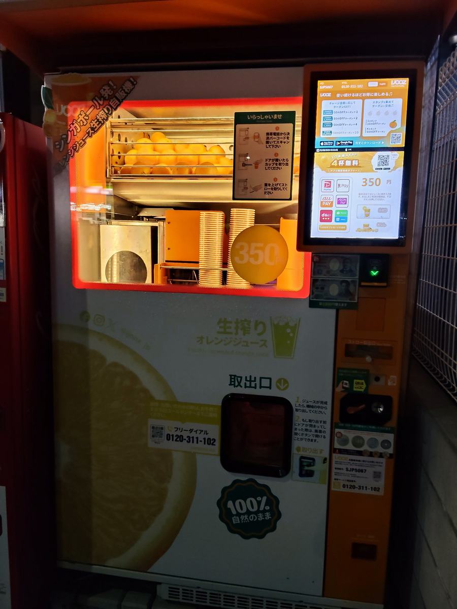 生搾りオレンジジュースの自販機。中に積まれたオレンジがライトに照らされている。