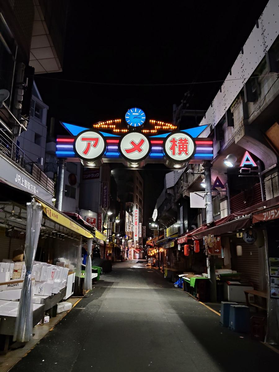 午前3時11分のアメ横のゲート。シャッターの下りた無人の通りの上で、看板と時計だけが光っている。