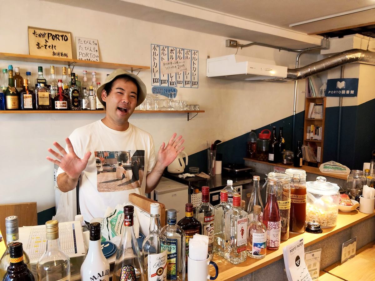 カウンターの内側で両手を広げる、バケットハットの喜屋武さん。背後の壁にはポルトカレーの手書き短冊がずらりと並ぶ。