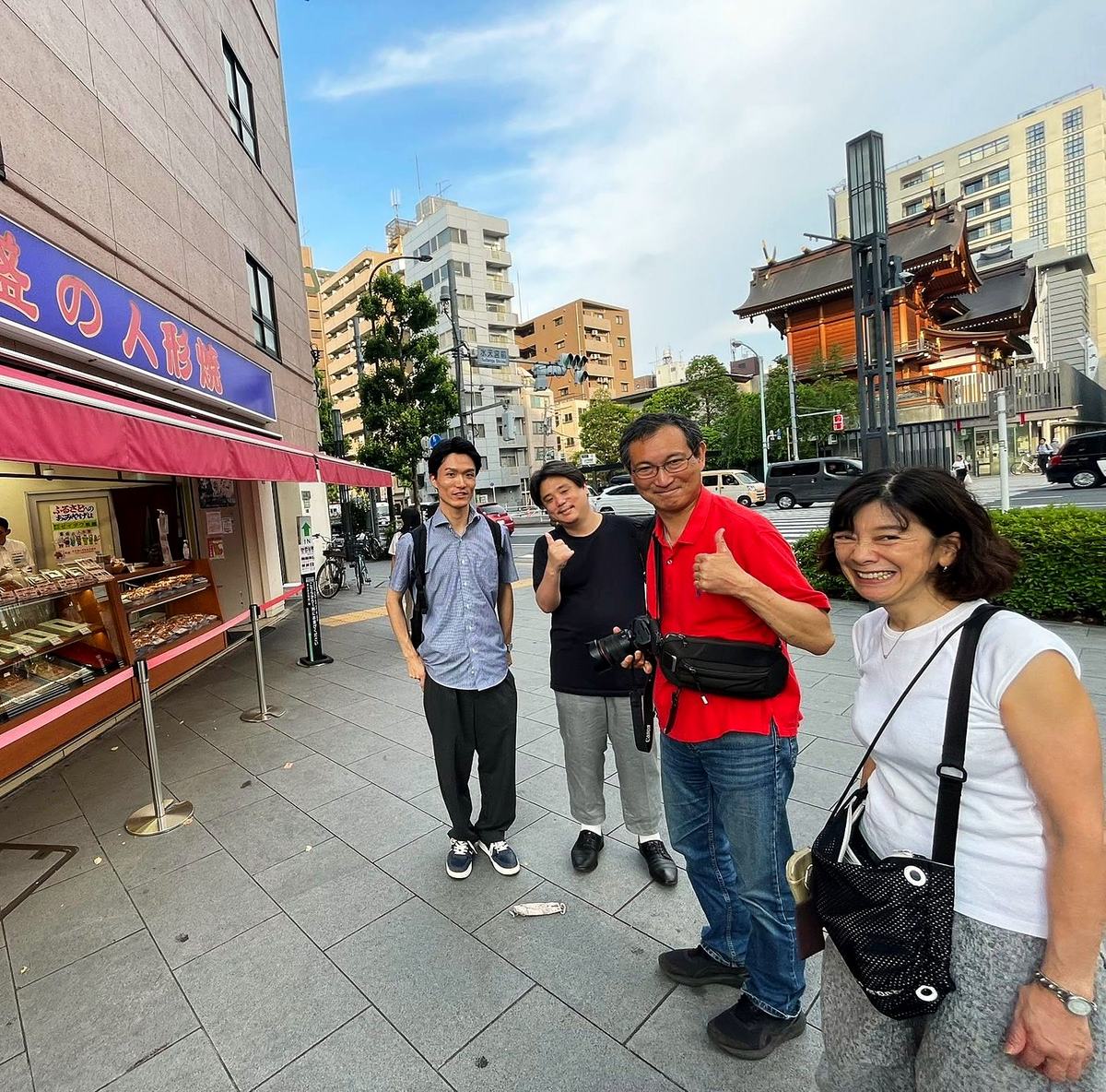 人形町の人形焼店の前に立つ四人。奥に水天宮の社殿。