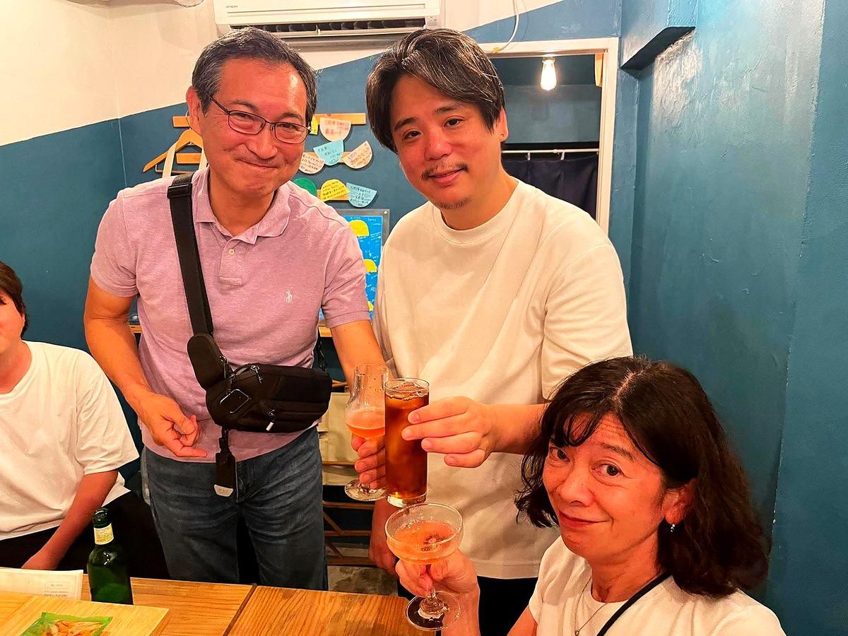 PORTOの店内、紺の壁の前で三人がグラスを掲げて乾杯している。