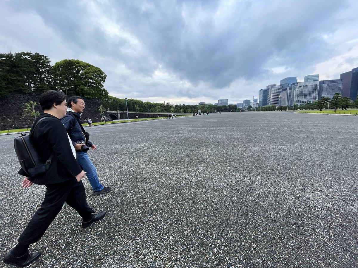 曇り空の皇居前広場を歩くふたり。ひとりは一眼レフを手に提げている。遠くに丸の内のビル群。