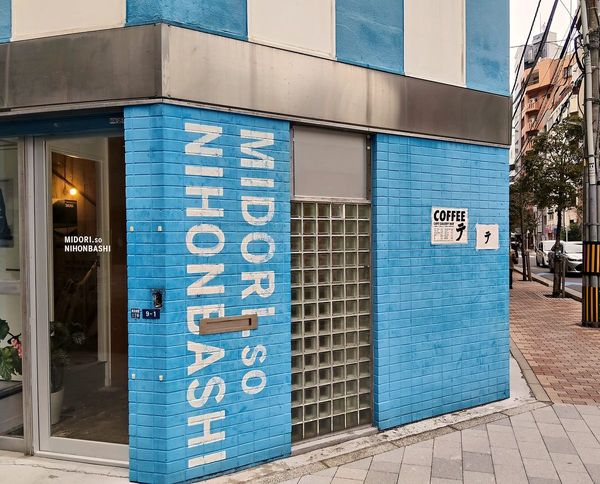 朝のテ。青いタイルのビルの角、ガラスブロックの壁に小さな「テ」の貼り紙。