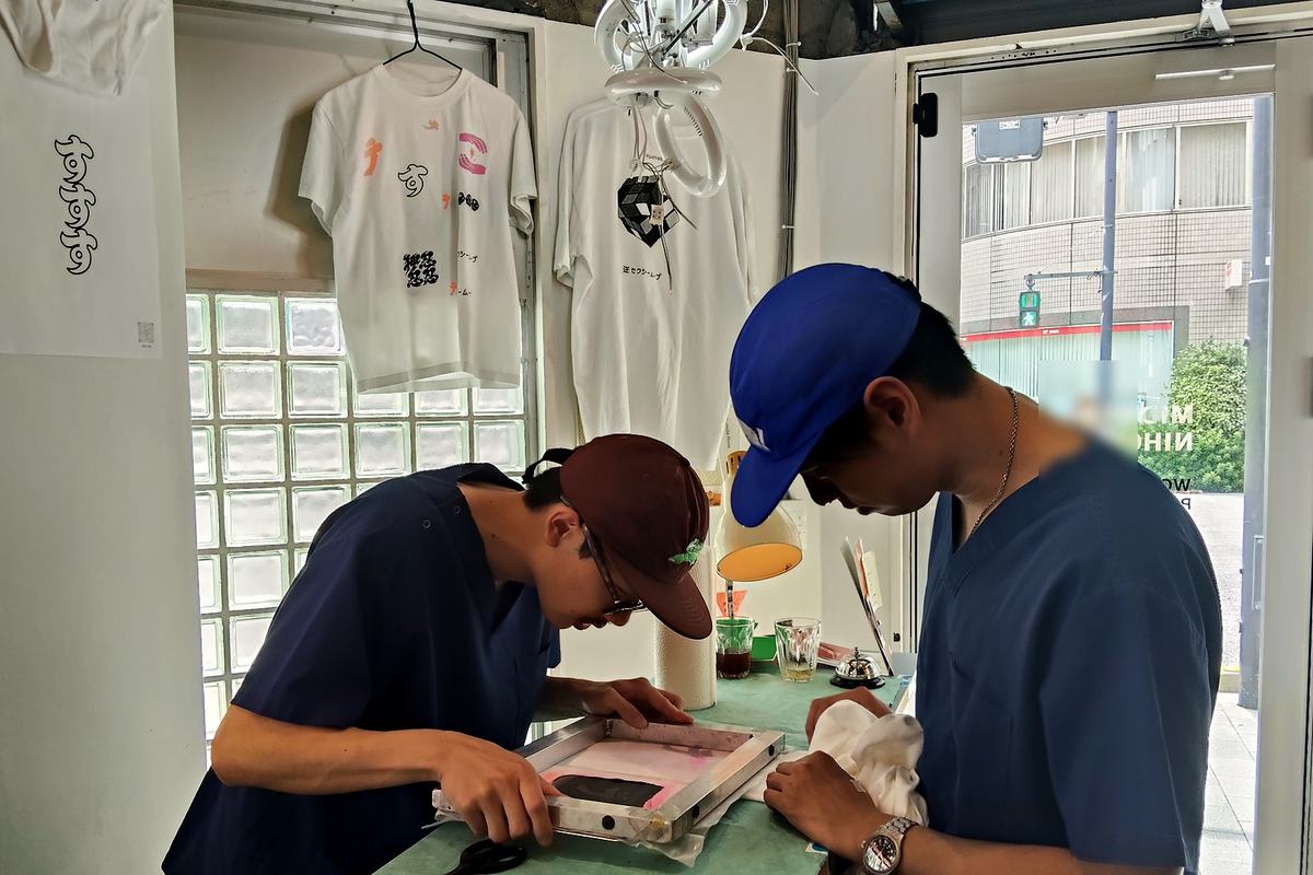 作業台の上のスクリーンを覗き込む二人。奥にはプリント済みのTシャツが吊るされている。
