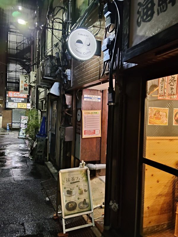 雨あがりの夜の小道。濡れた路面に光が映り、奥に丸い「Cafe & Bar Terra Cotta」の看板が白く灯っている。