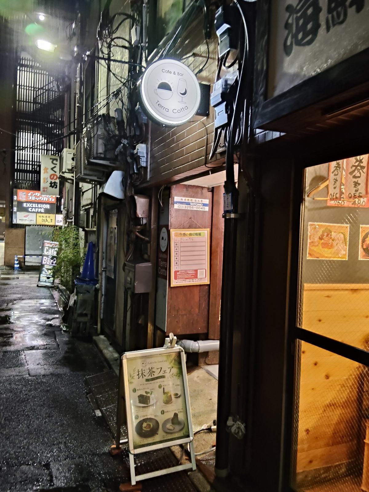 雨あがりの夜の小道。濡れた路面に光が映り、奥に丸い「Cafe & Bar Terra Cotta」の看板が白く灯っている。