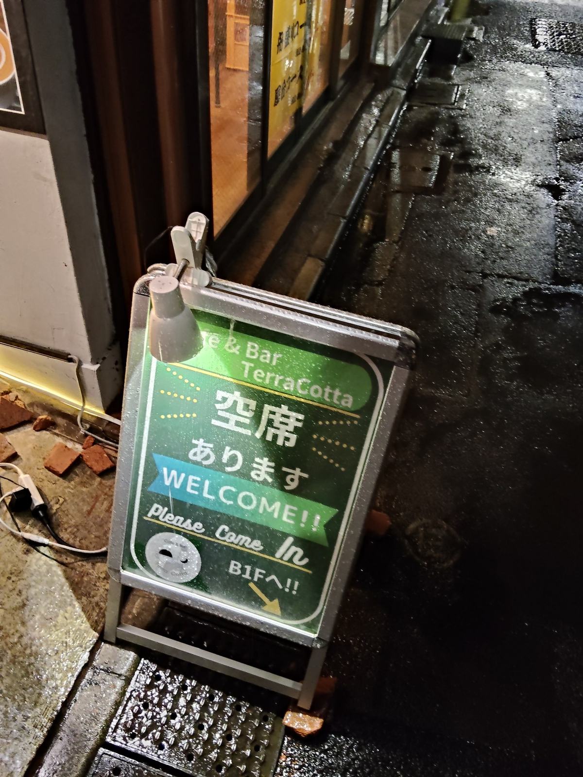 濡れた路上に置かれた緑の立て看板。「空席あります WELCOME!!」の文字がスポットライトで照らされている。