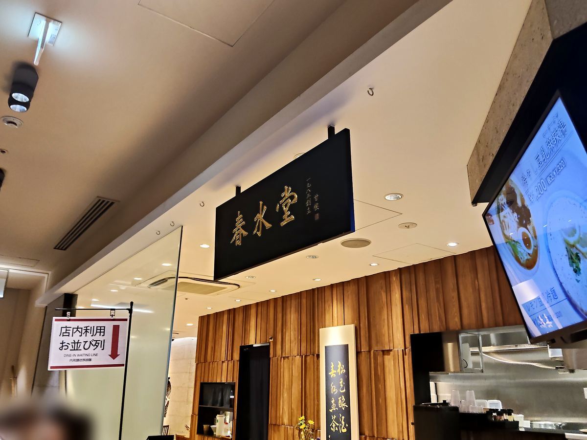 春水堂の店先。黒地に金の看板が掛かり、木目の壁が続いている。